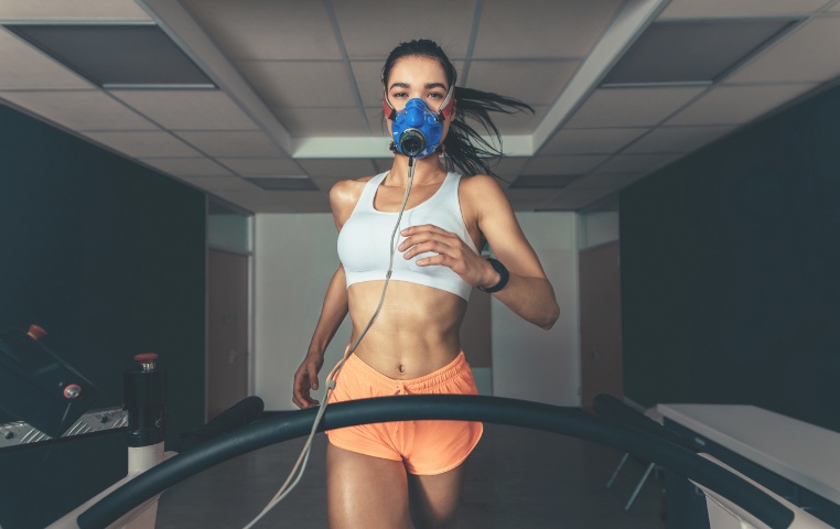 A woman running on a treadmill while wearing a VO2 max testing mask, in a laboratory setting, indicating the measurement of her cardiovascular fitness during intense exercise.