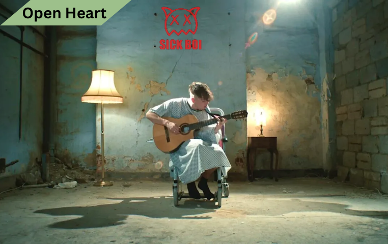 Ren Gill, dressed in a hospital gown, plays an acoustic guitar while seated in a wheelchair in a dimly lit, worn-out room with peeling walls. Above him, the Sick Boi logo is displayed in red.